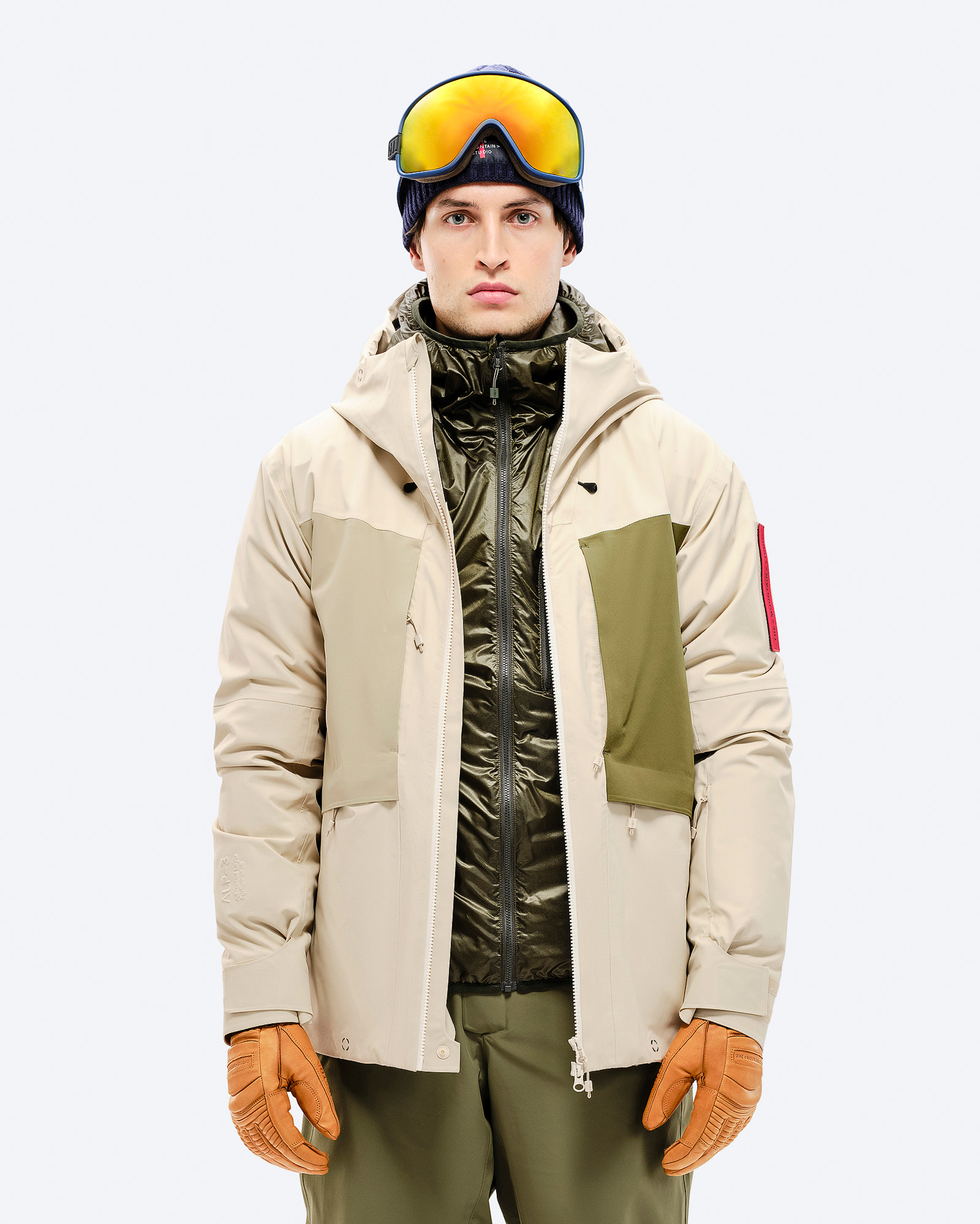 Skifahrer in Hybridjacke Mann mit Skibrille und Mütze, trägt beige 3L Hybrid Shell Jacke mit grünen Akzenten und Lederhandschuhen.