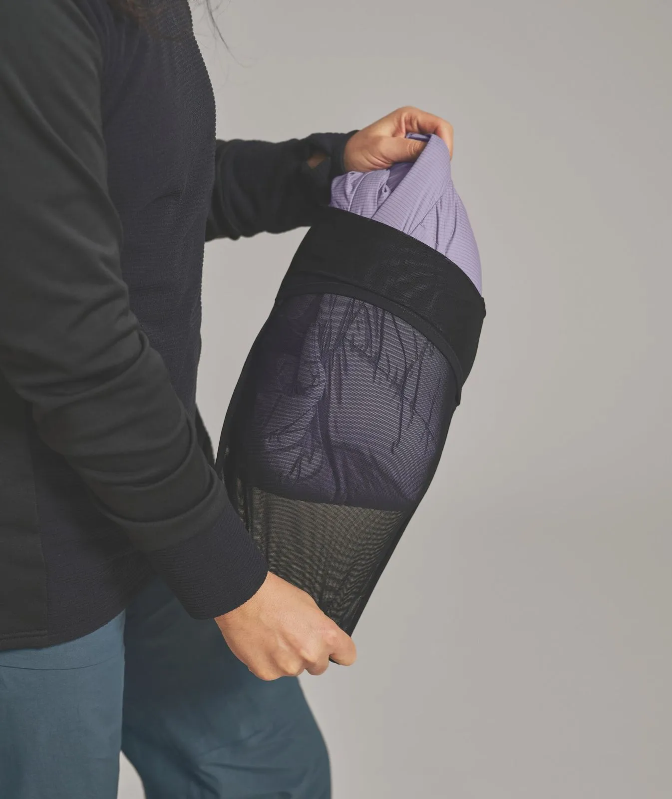 Person packt lila Kleidung in einen schwarzen Netzbeutel. Die Person trägt einen schwarzen Pullover und grüne Hose.