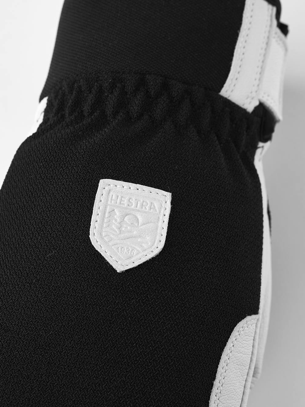 Close-up of a black glove with white leather accent and a Hestra logo.