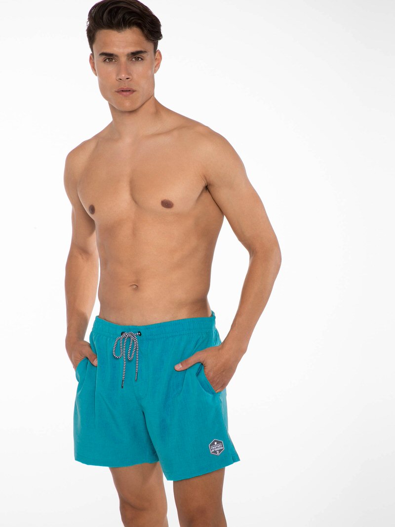 Mann mit blauen Protest Davey Sea Bay Badeshorts, Hände in den Taschen.