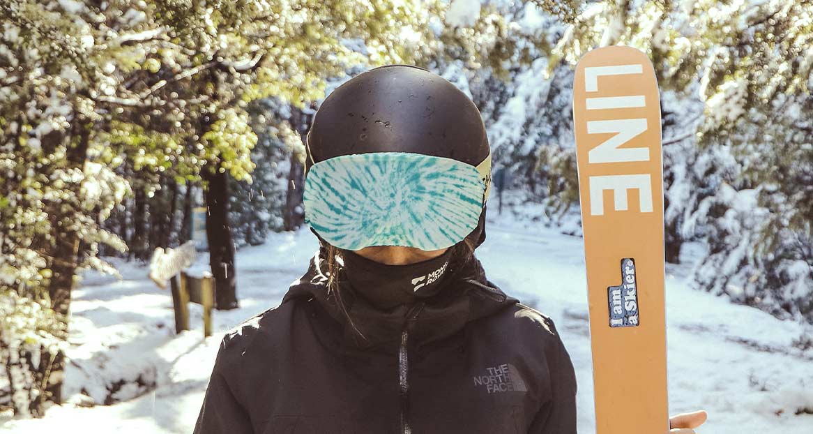 Skifahrer im Schnee Skifahrer mit Helm und Gogglesoc steht im Schnee. Eine Skispitze mit dem Wort "LINE" ist sichtbar. Bäume mit Schnee im Hintergrund.