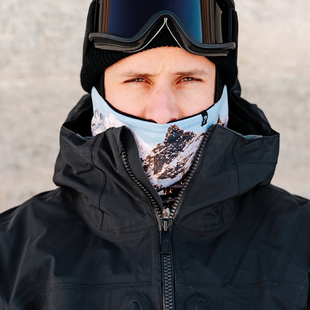 Skifahrer mit Neckwarmer Porträt einer Person mit schwarzer Mütze, Skibrille und "The Mask Black" Neckwarmer mit Bergmotiv unter einer schwarzen Jacke.