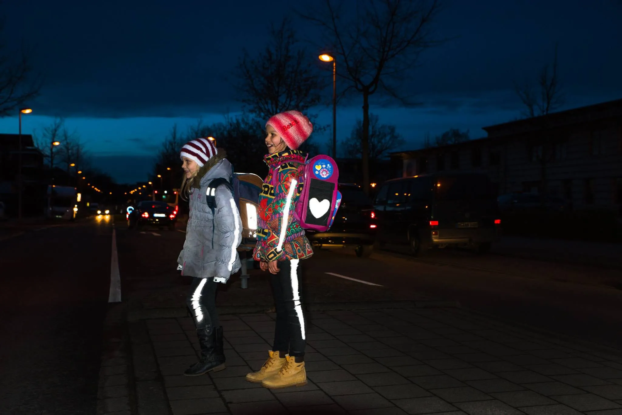 Zwei Mädchen mit reflektierender Kleidung stehen nachts an einer dunklen Straße. Autos sind im Hintergrund zu sehen.
