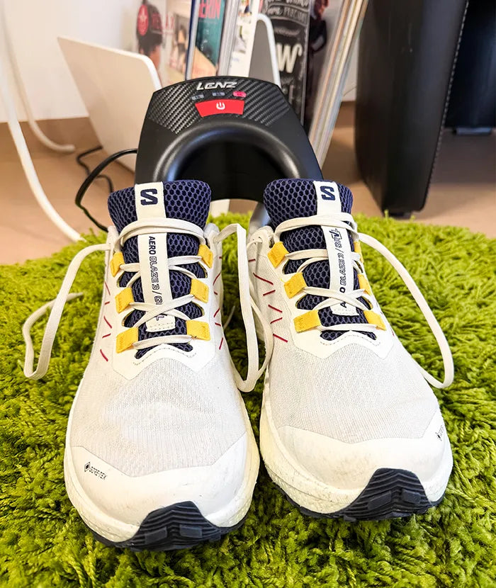 A pair of white running shoes with yellow accents on a green, shaggy carpet against a blurred background. The laces are loosely tied.