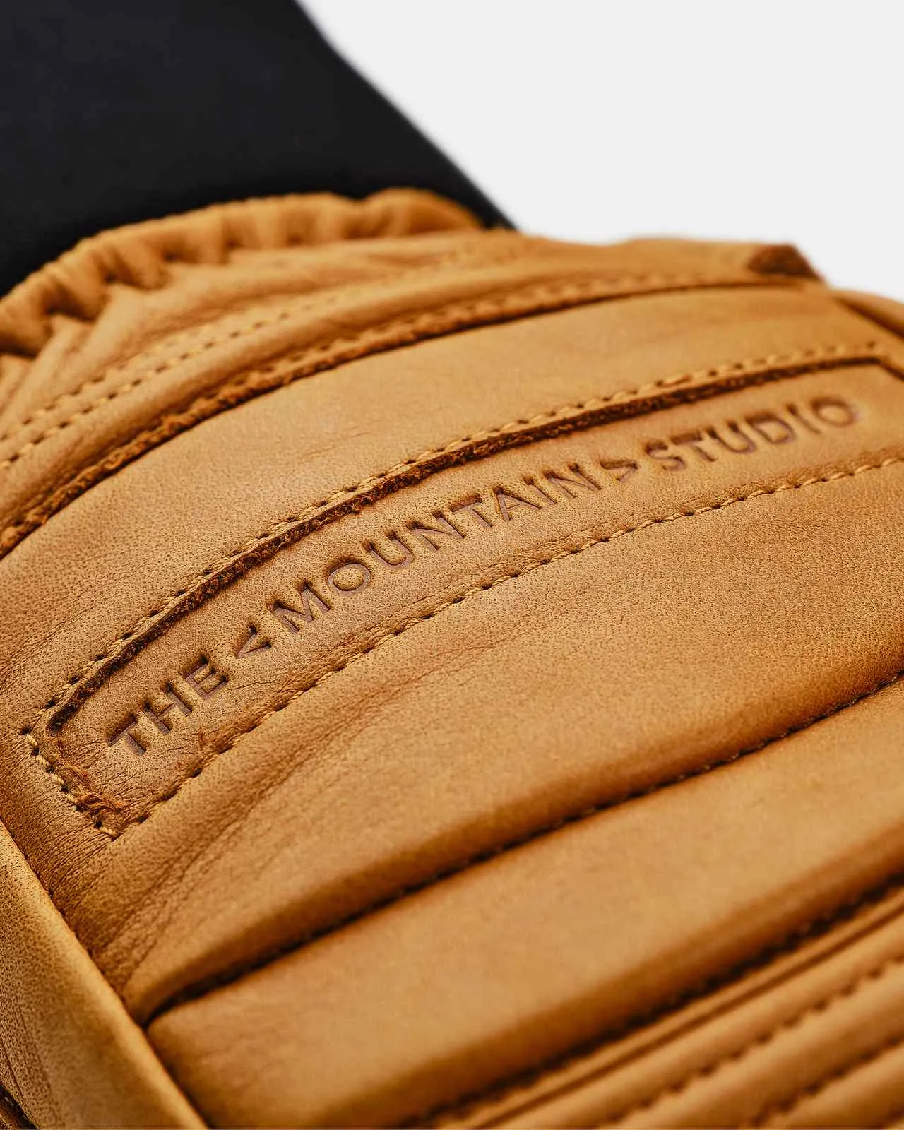 Nahaufnahme eines Lederhandschuhs in Sandfarbe mit "THE MOUNTAINS STUDIO"-Prägung und Steppdetails.