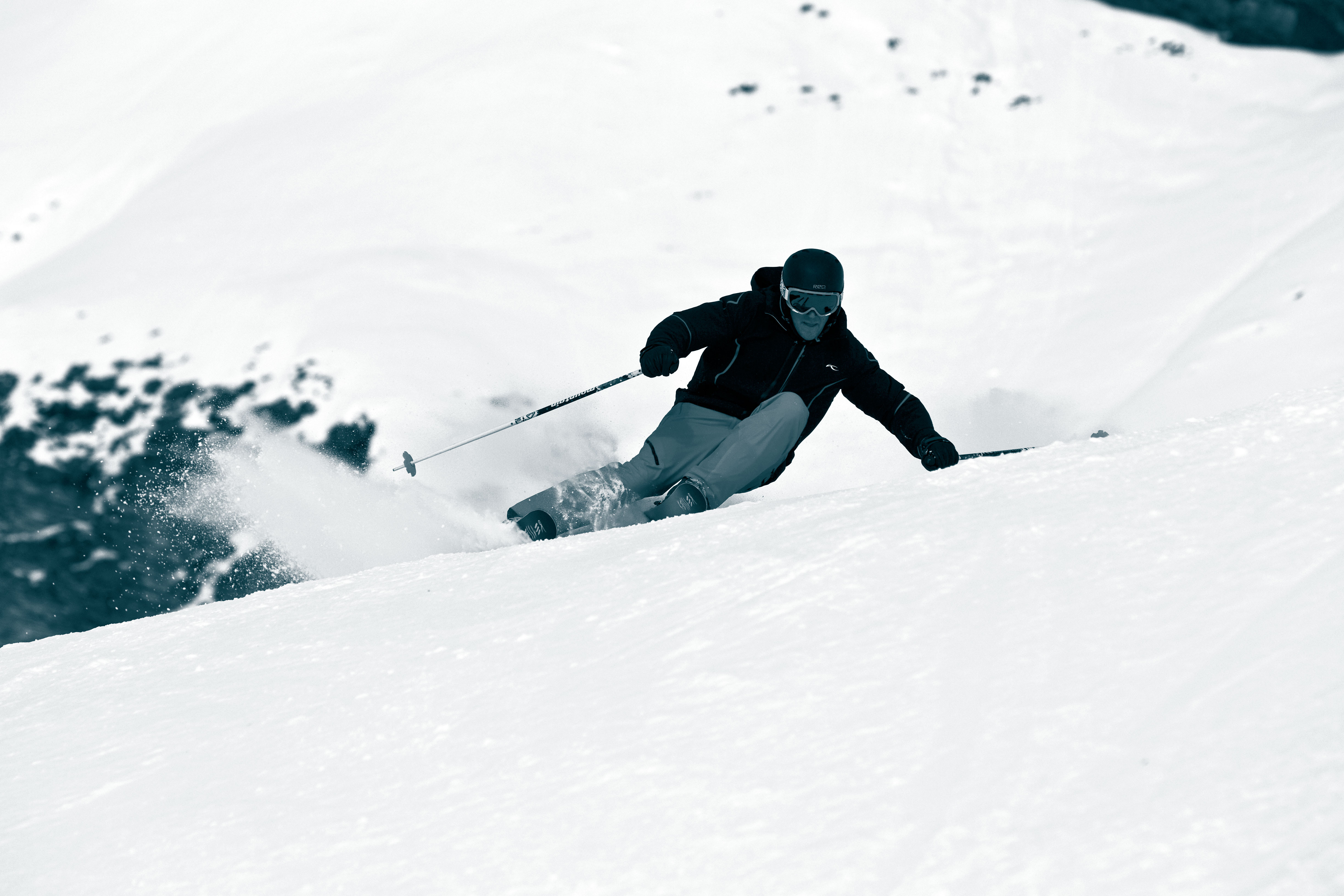 Skifahrer im Schnee Skifahrer in voller Montur fährt einen schneebedeckten Hang hinunter, der Schnee spritzt. Berglandschaft im Hintergrund.