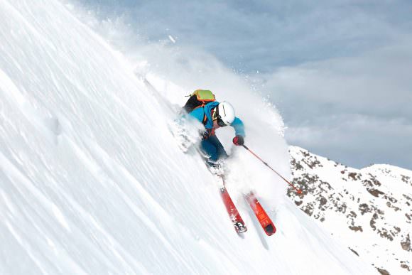 Skitourengeher mit Dynafit Hoji Free Ski in Action, Pulverschnee spritzt, Berglandschaft.