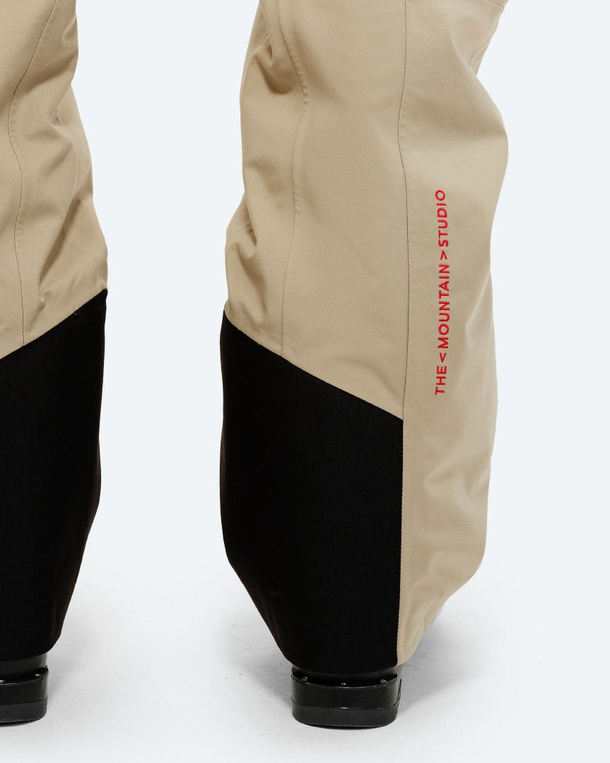 P-7 Ride Isolierte Hose Detailaufnahme: Untere Hälfte der P-7 Ride Insulated Pant in Beige mit schwarzem, diagonalen Farbblock und rotem Markenlogo.