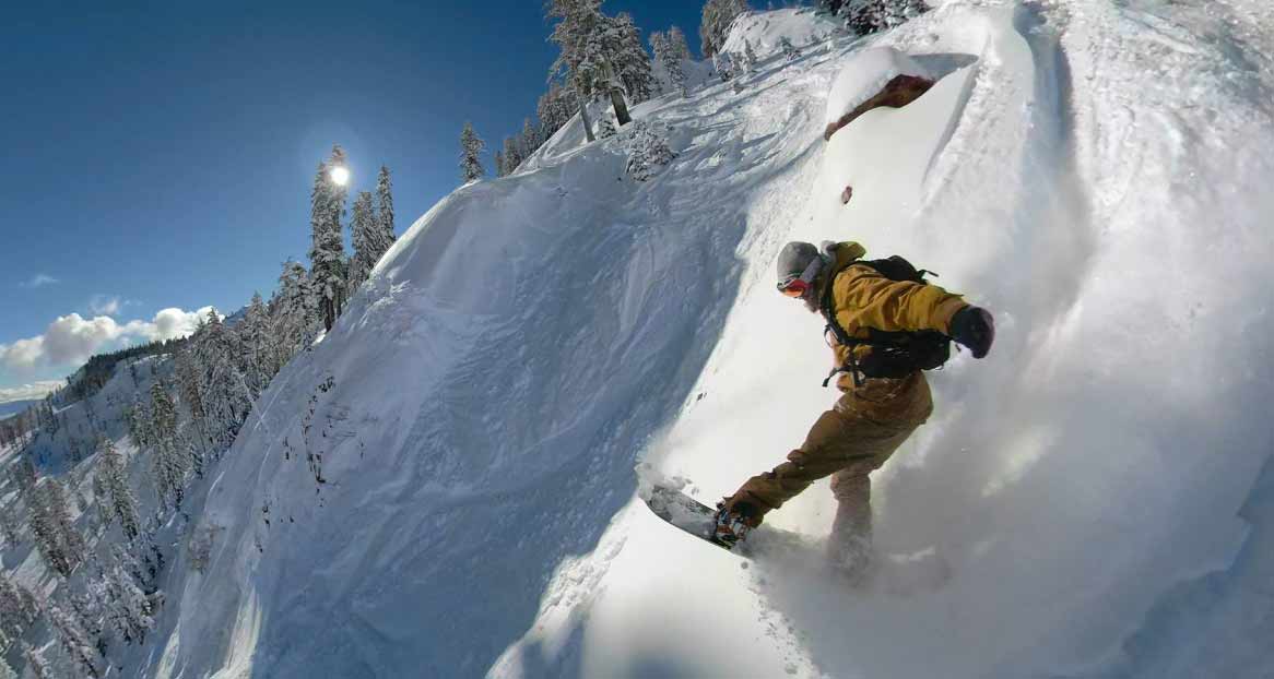 Snowboarder in the snow A snowboarder rides down a steep, snow-covered slope, surrounded by trees and under a blue sky.