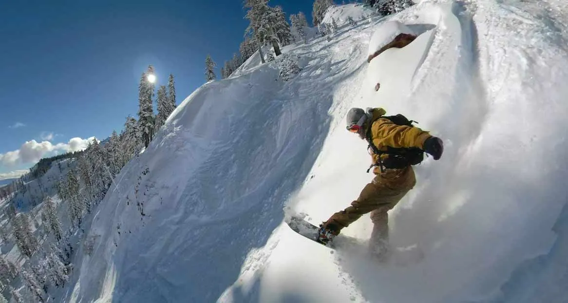 Ein Snowboarder fährt einen steilen, schneebedeckten Hang hinunter, umgeben von Bäumen und unter einem blauen Himmel.