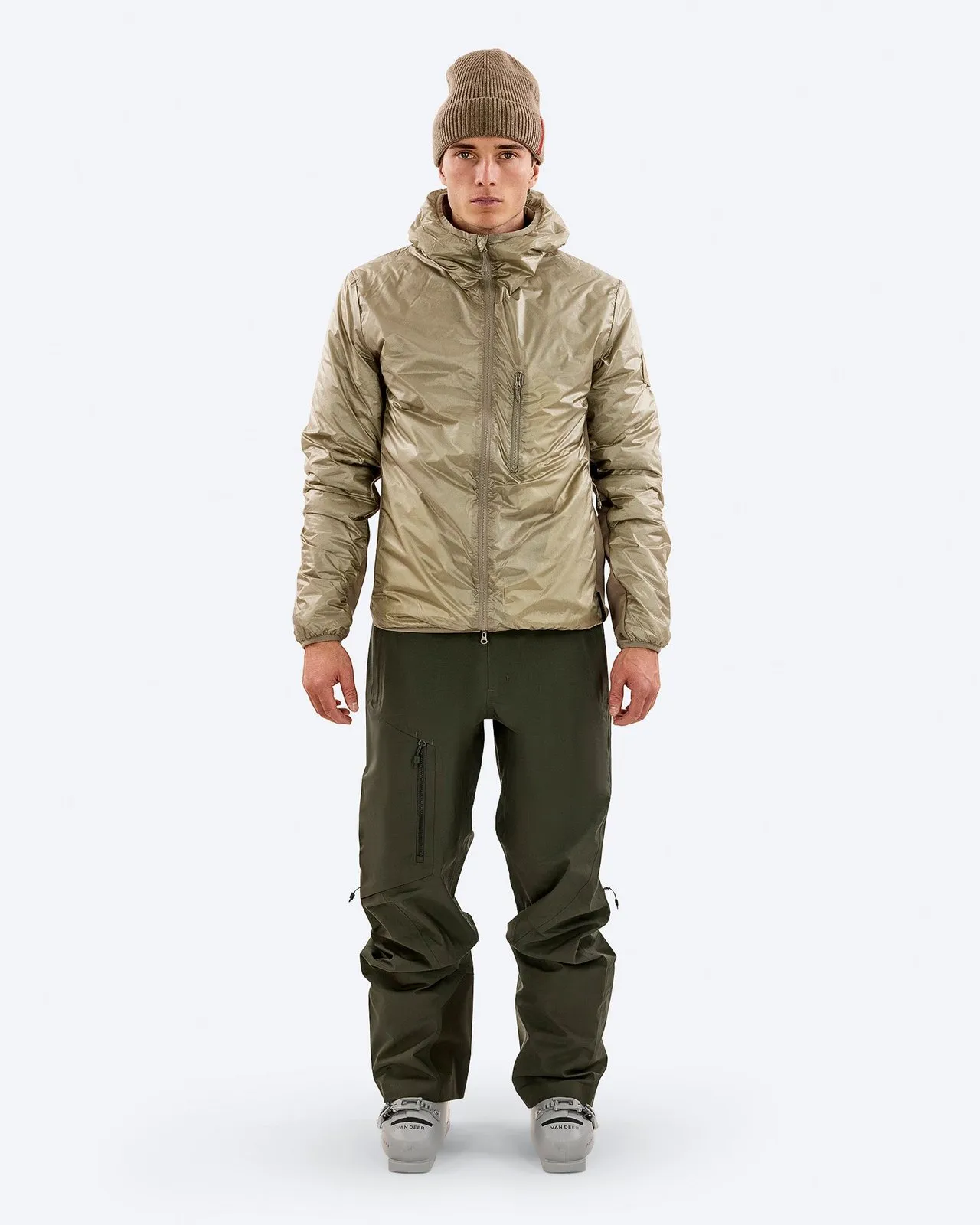 Man in outdoor clothing: Brown cap, shimmering jacket with hood, olive green pants and ski boots against a white background.
