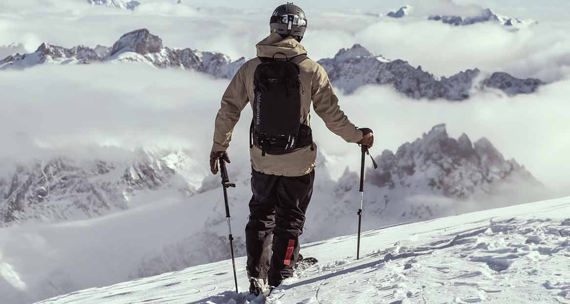 Skieur de dos sur une pente enneigée, avec des bâtons. En arrière-plan, hautes montagnes partiellement nuageuses.
