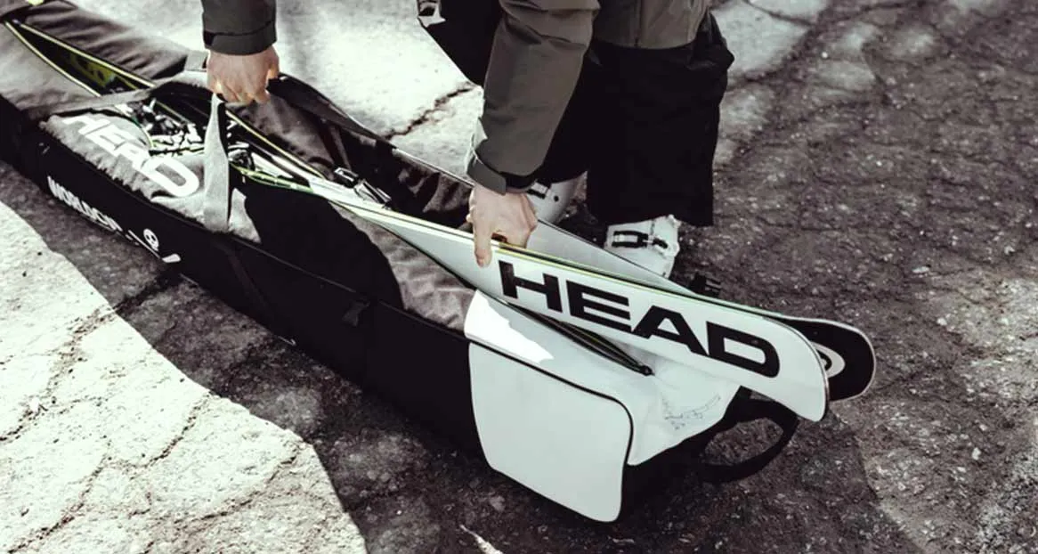 A person packs "Head" skis into a black ski bag. The bag is standing on a rough surface. Ski boots are visible in the background.