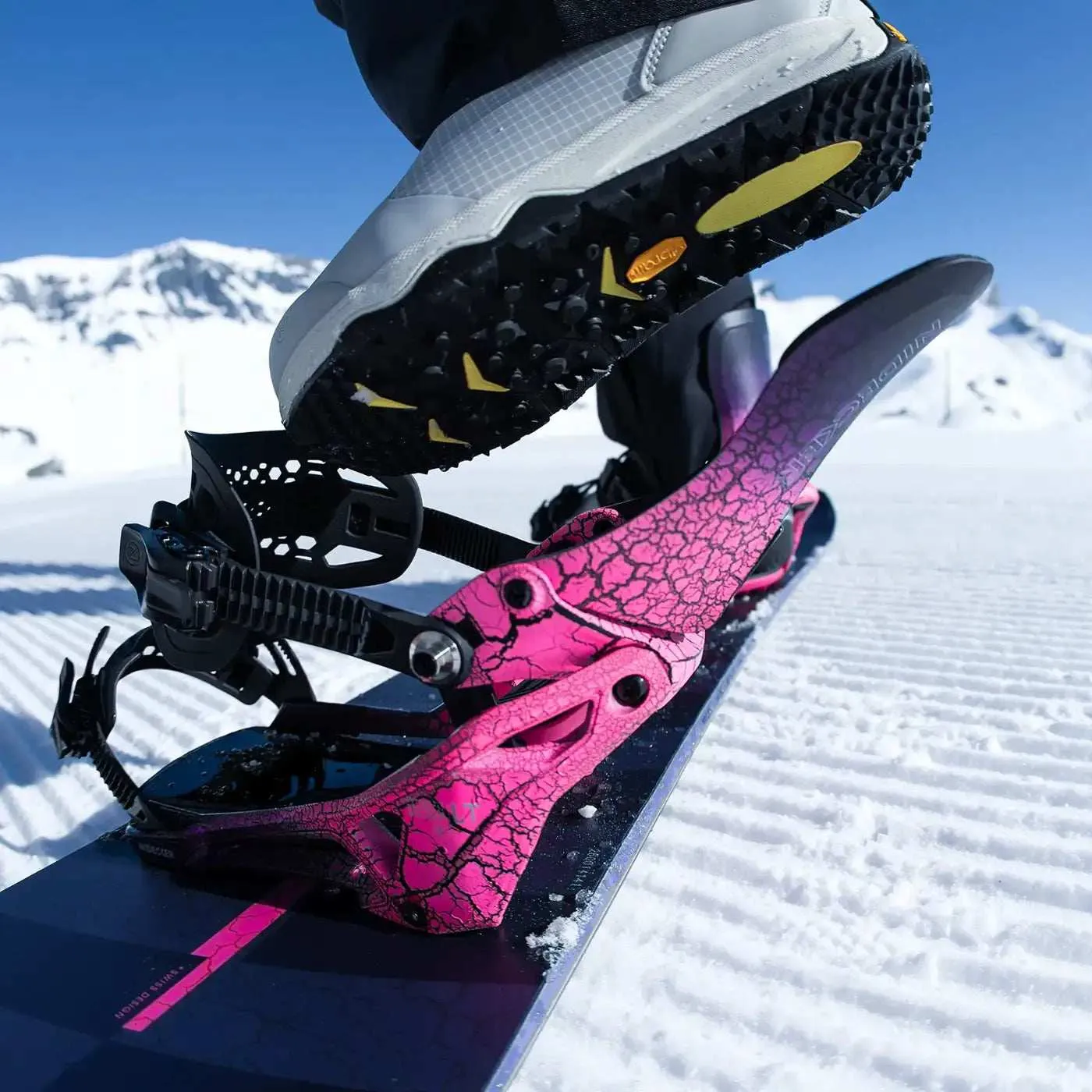 Primo piano di uno snowboard con attacchi rosa e uno scarpone grigio con suola nera su un piede. Sullo sfondo montagne innevate.