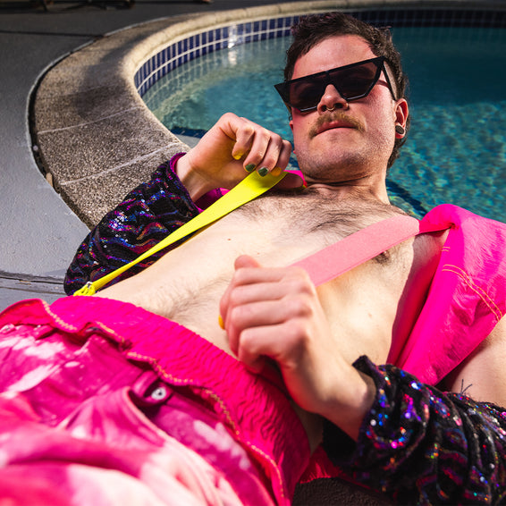 Mann mit Sonnenbrille und Oberkörper frei, mit Hosenträgern und pinker Hose, am Pool liegend.