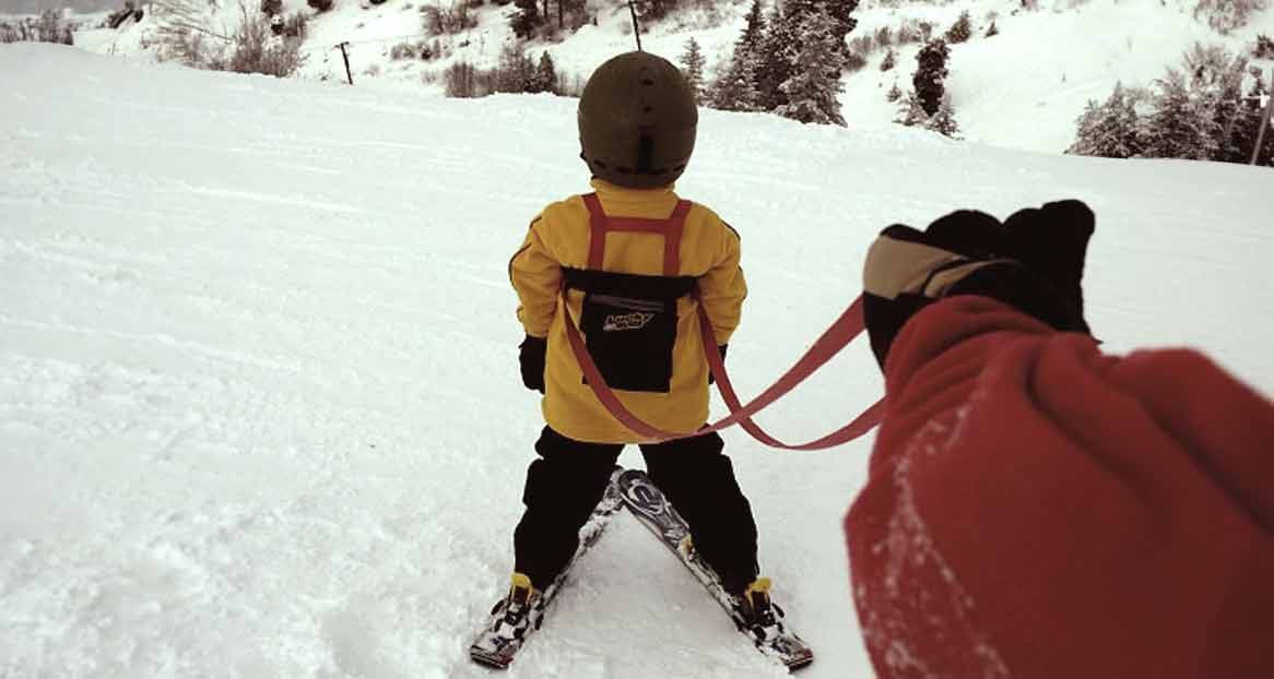 Enfant portant un casque de ski et une veste jaune, tiré sur la neige par une personne portant un gant rouge et tenant une corde rouge.