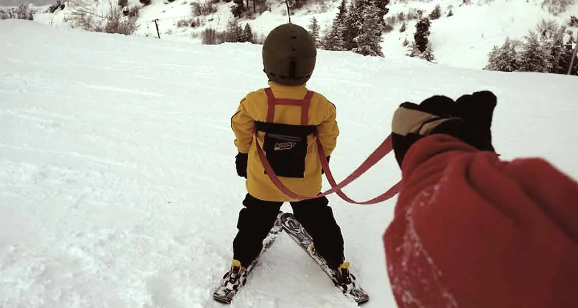 Child with ski helmet and yellow jacket being pulled on snow by a person with a red glove on a red leash.