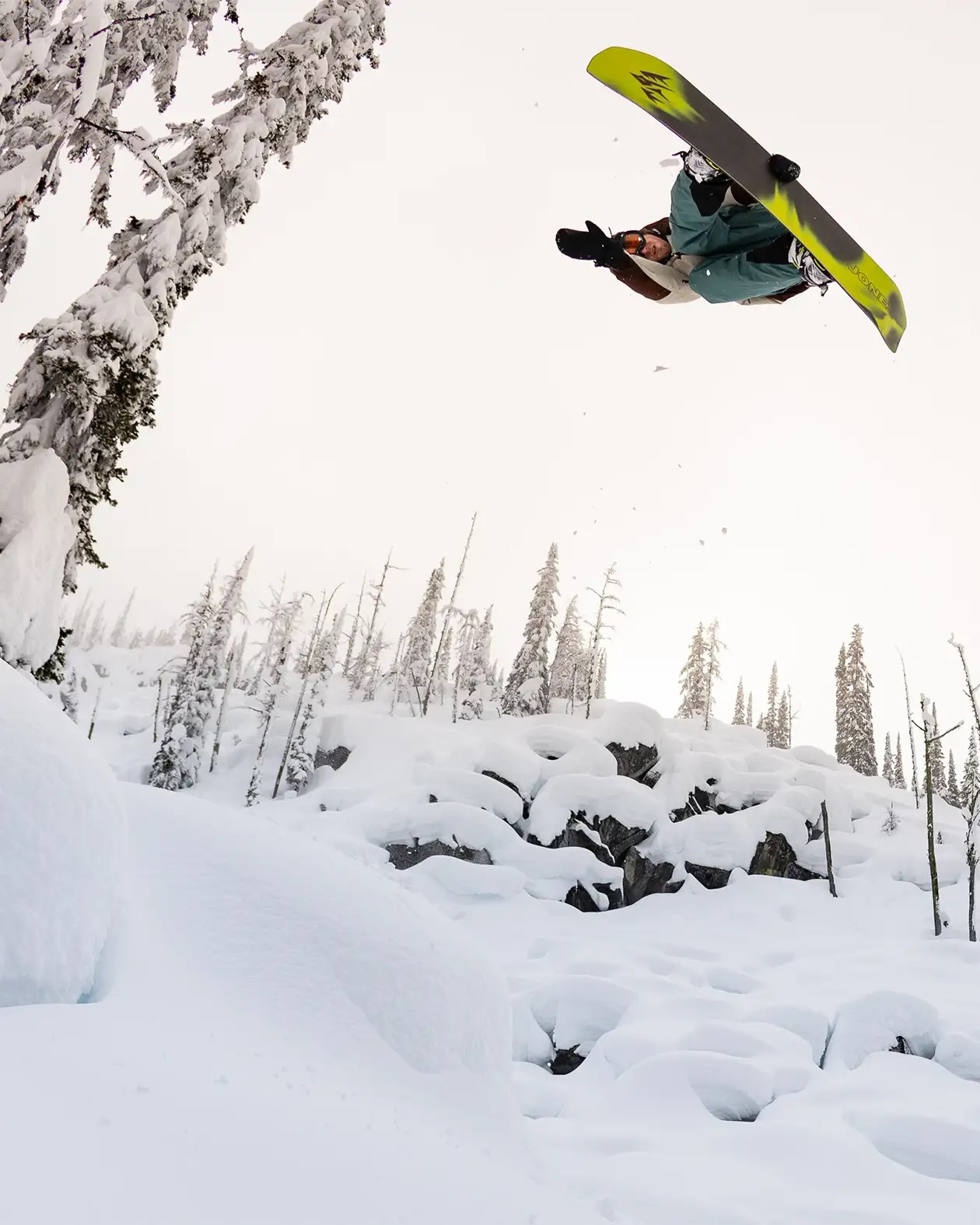 Snowboarder macht einen Trick in der Luft vor einer verschneiten Landschaft mit Bäumen.