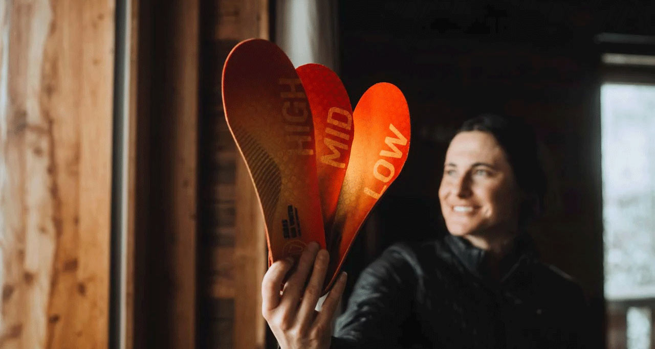 Une femme souriante tient dans sa main trois semelles intérieures orange, étiquetées "HIGH", "MID" et "LOW". En arrière-plan, on peut voir un mur en bois.