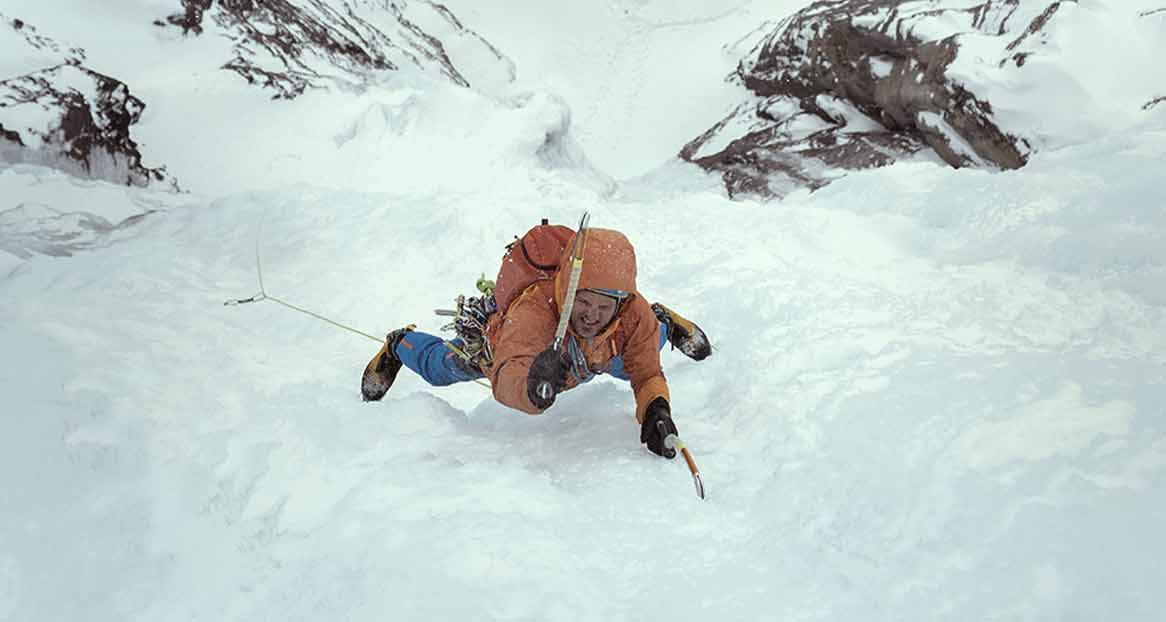 Un alpiniste escalade une paroi de montagne enneigée. Il porte une veste orange et un pantalon bleu et est équipé de crampons.