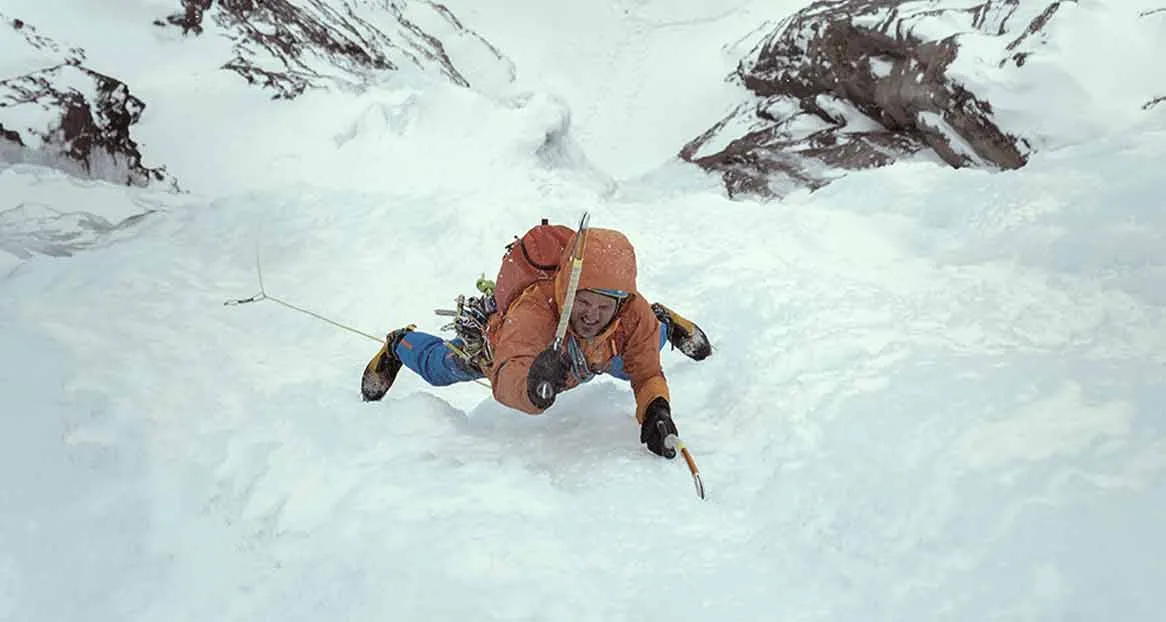 A mountaineer climbs up a snowy mountain face. He is wearing an orange jacket and blue trousers and has crampons on.