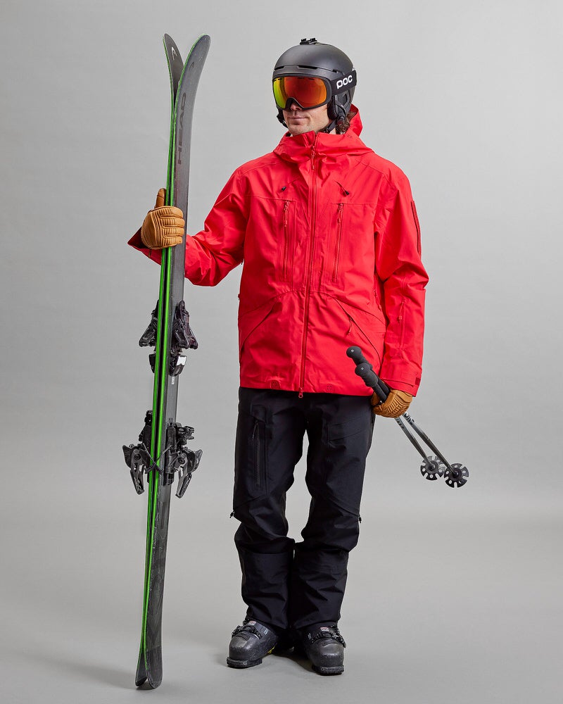 Skifahrer mit Ausrüstung Person in Skikleidung, rote Jacke, schwarze Y-1 HD GORE-TEX Pro Hose, Helm, Skibrille, Skier und Stöcke.