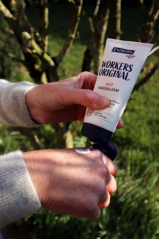 Enzborn Handbalsam Anwendung Eine Person trägt Enzborn Workers Original Handbalsam auf den Handrücken auf. Im Hintergrund sind Bäume und Gras zu sehen.
