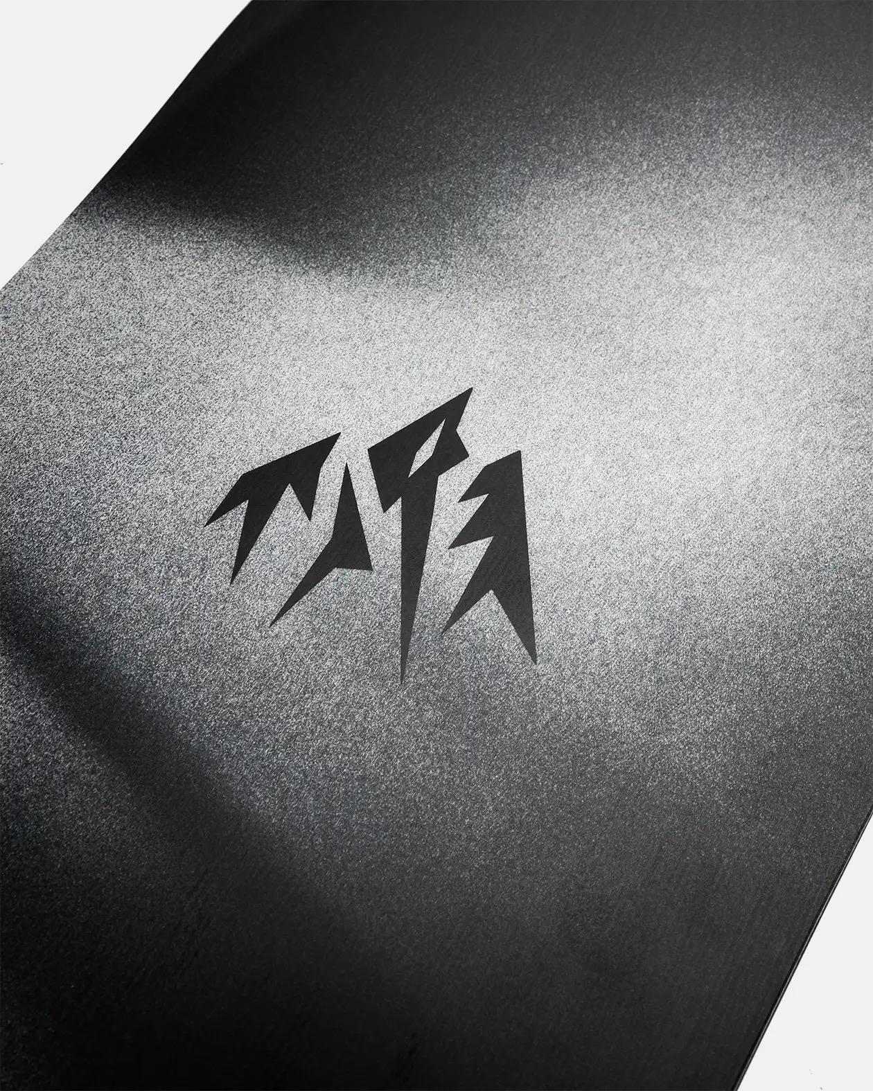 Auf einer hellgrauen, leicht körnigen Oberfläche ist ein dreiteiliges, zackiges, schwarzes Logo abgebildet. Der Hintergrund ist zum oberen Rand hin dunkler.