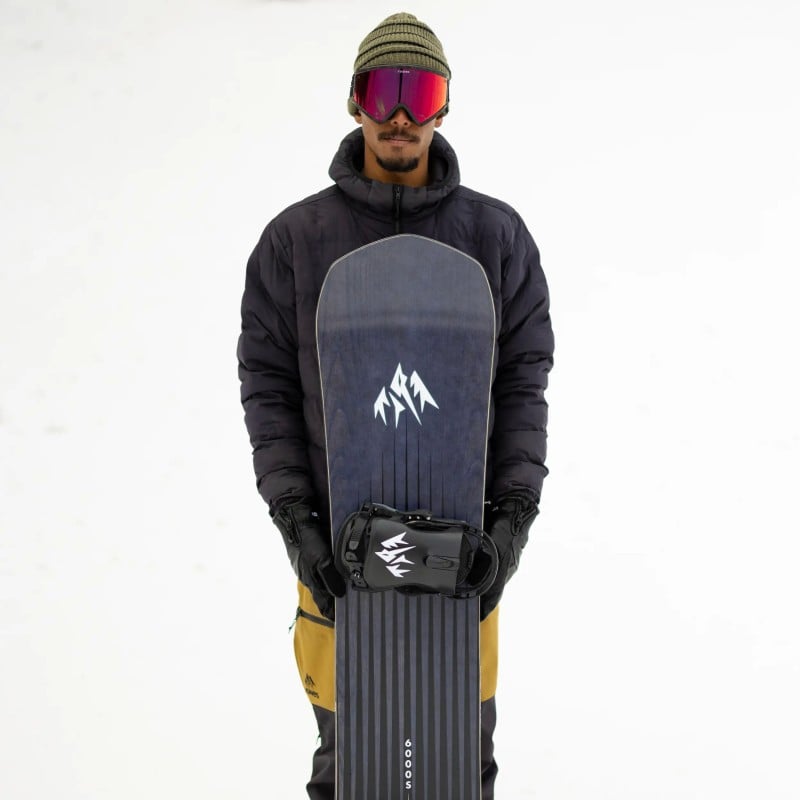 Jones Freecarver Snowboard Person hält ein Jones Freecarver Snowboard mit Bindung vor sich. Er trägt Mütze, Skibrille, Jacke und Handschuhe.