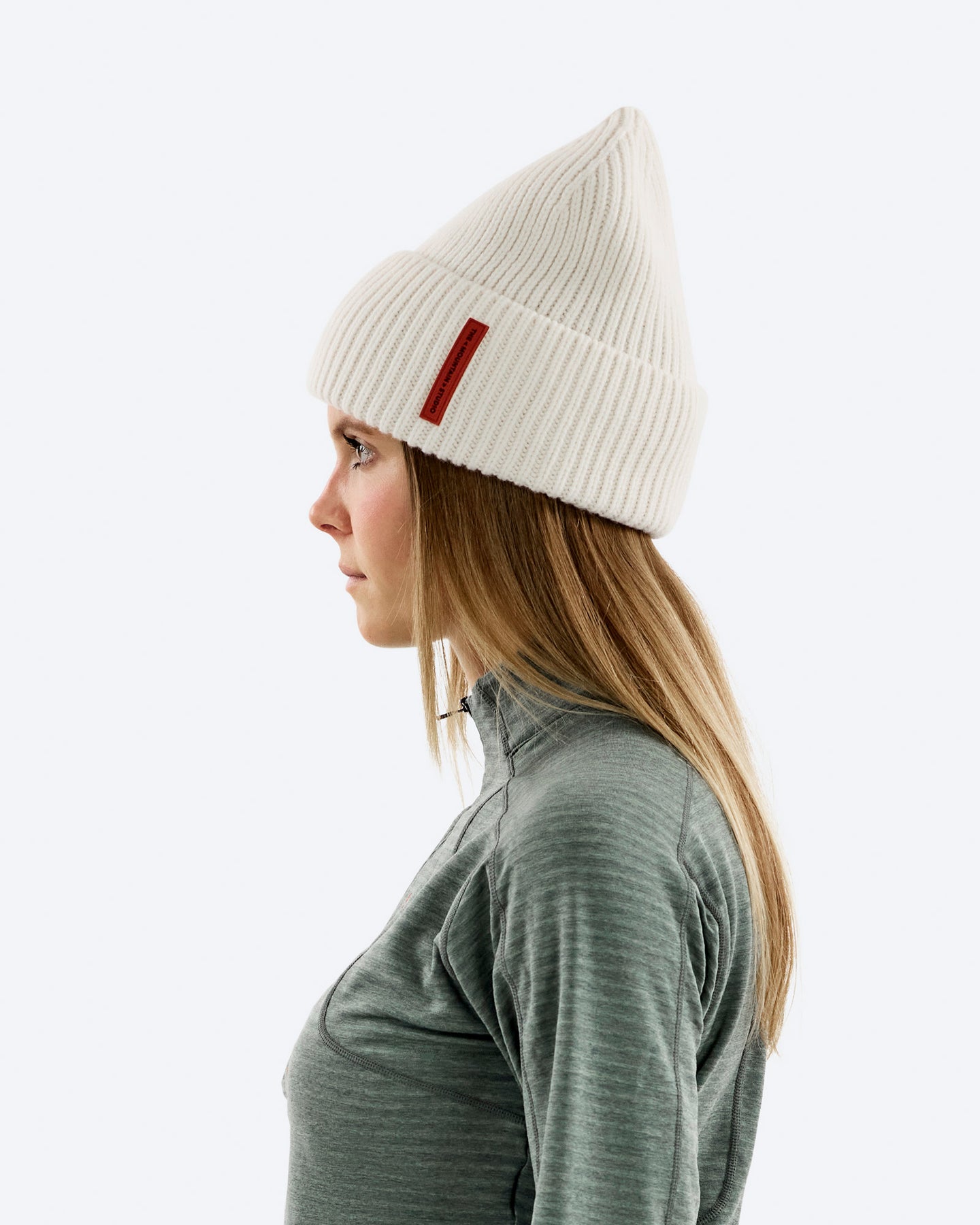 Profil einer Frau mit blonden Haaren, die eine gerippte weiße Mütze mit rotem Etikett und ein graues Oberteil trägt.