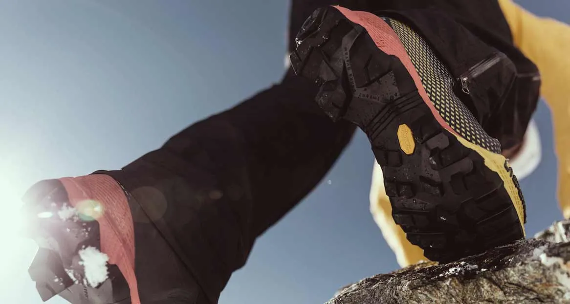 Close-up of a hiking boot with a coarse tread on a rock against a blue sky. The tread is black, with yellow and red accents.