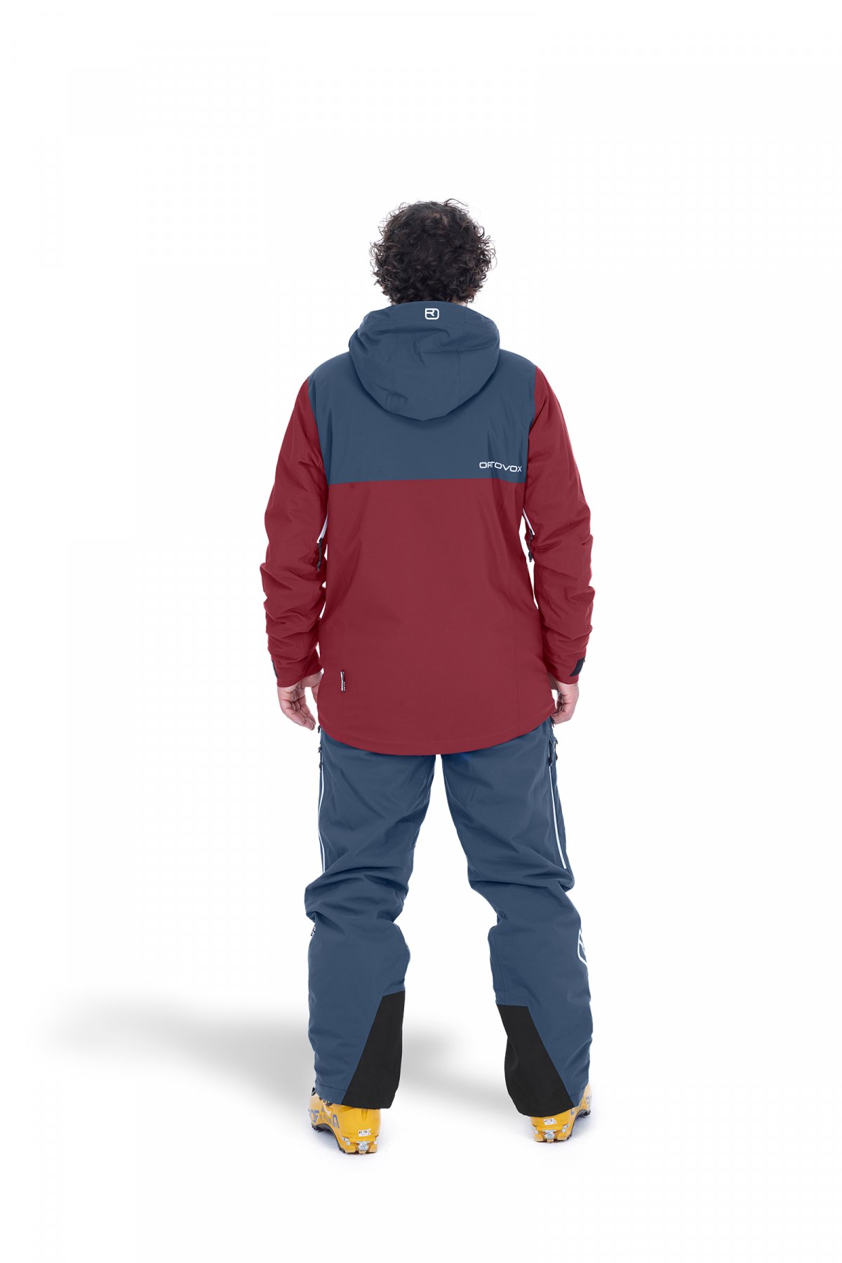 Tenue Andermatt noir acier Personne de dos avec veste et pantalon Andermatt en Black Steel. La veste est rouge et bleue, le pantalon est bleu.