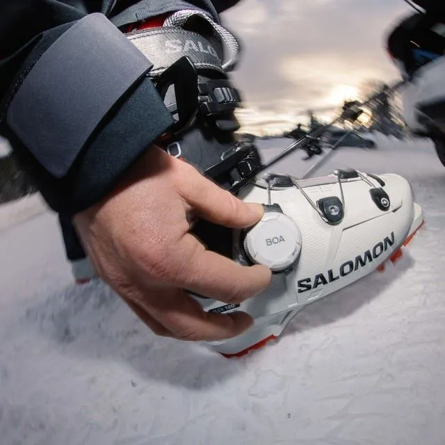 Gros plan : la main actionne la fermeture rotative BOA de la chaussure de ski blanche Salomon S/PRO SUPRA BOA 120 dans la neige.
