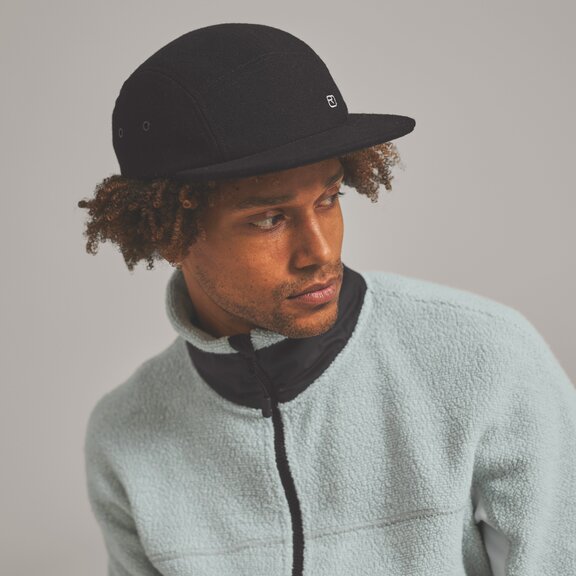 Ein Mann mit lockigem Haar trägt eine schwarze Kappe und eine hellblaue Fleecejacke. Er schaut zur Seite.