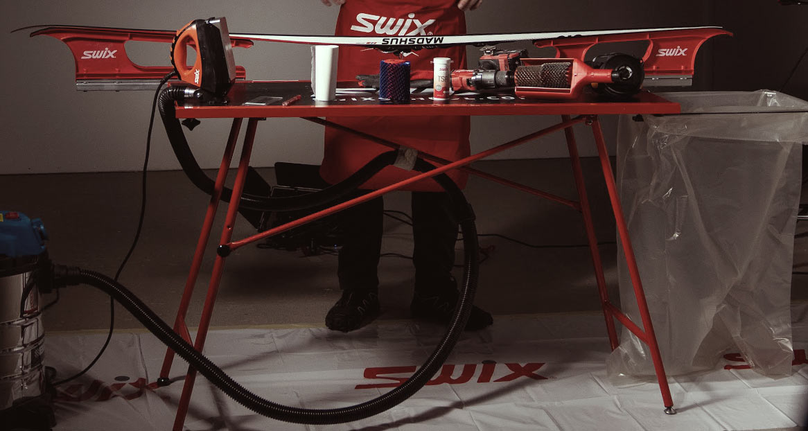 Skipaar mit Wachsen Zubehör Ein Skipaar auf einem Wachstisch mit Geräten zum Wachsen, inklusive Bügeleisen und Bürsten. Eine Person steht dahinter, trägt eine rote Swix-Schürze.