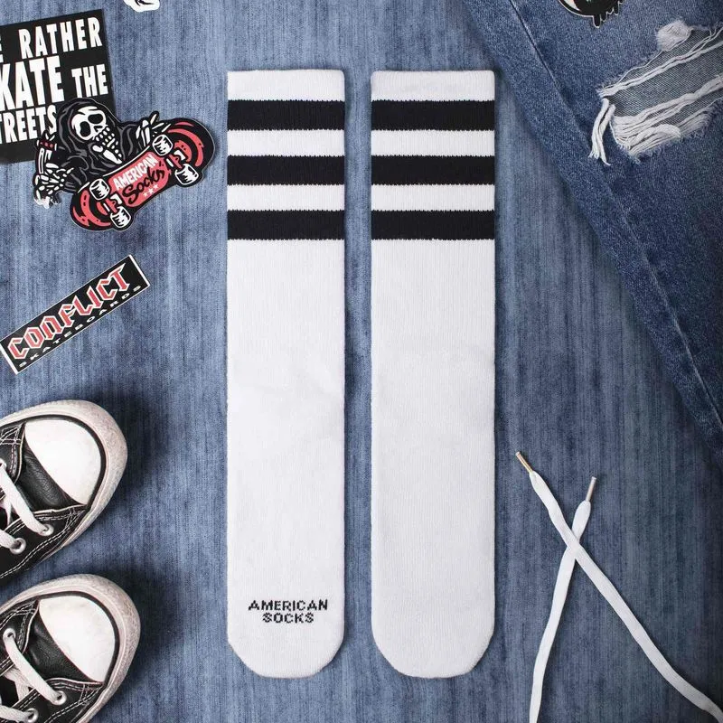 Two white mid high socks with black stripes, surrounded by jeans, shoes and stickers.