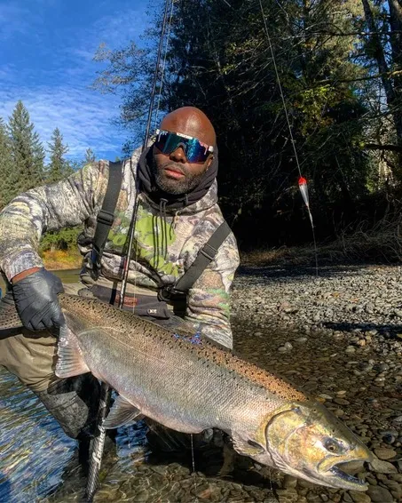 Uomo con occhiali da sole polarizzati che tiene in mano un grosso pesce nel fiume, in un ambiente boscoso.