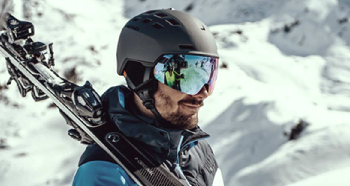 Skifahrer in den Bergen Mann mit Skihelm und reflektierender Brille, Skier über der Schulter, im Hintergrund verschneite Berge.