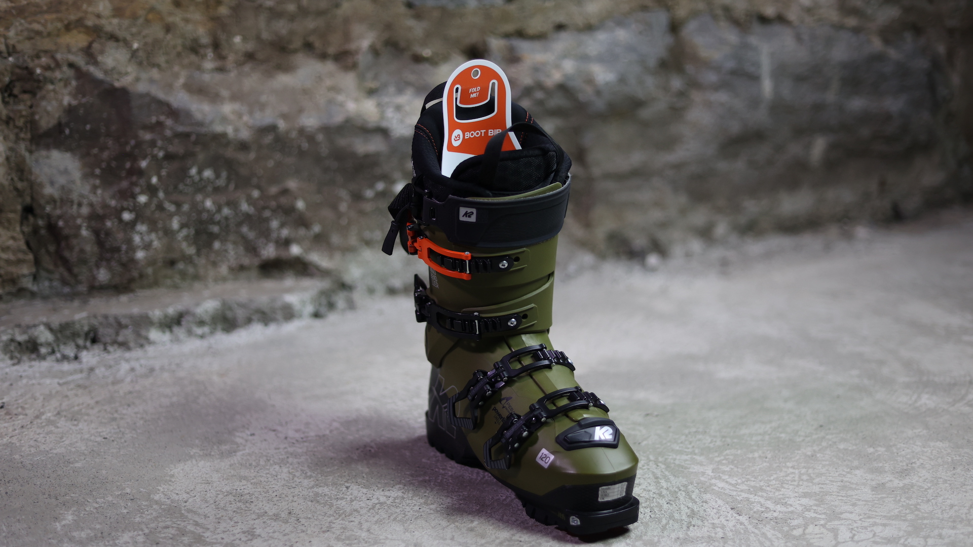 Olivgrüner K2 Skischuh mit Schnallen und orangenen Akzenten steht vor einer Steinwand. Innenliegend ist ein Größenhinweis.