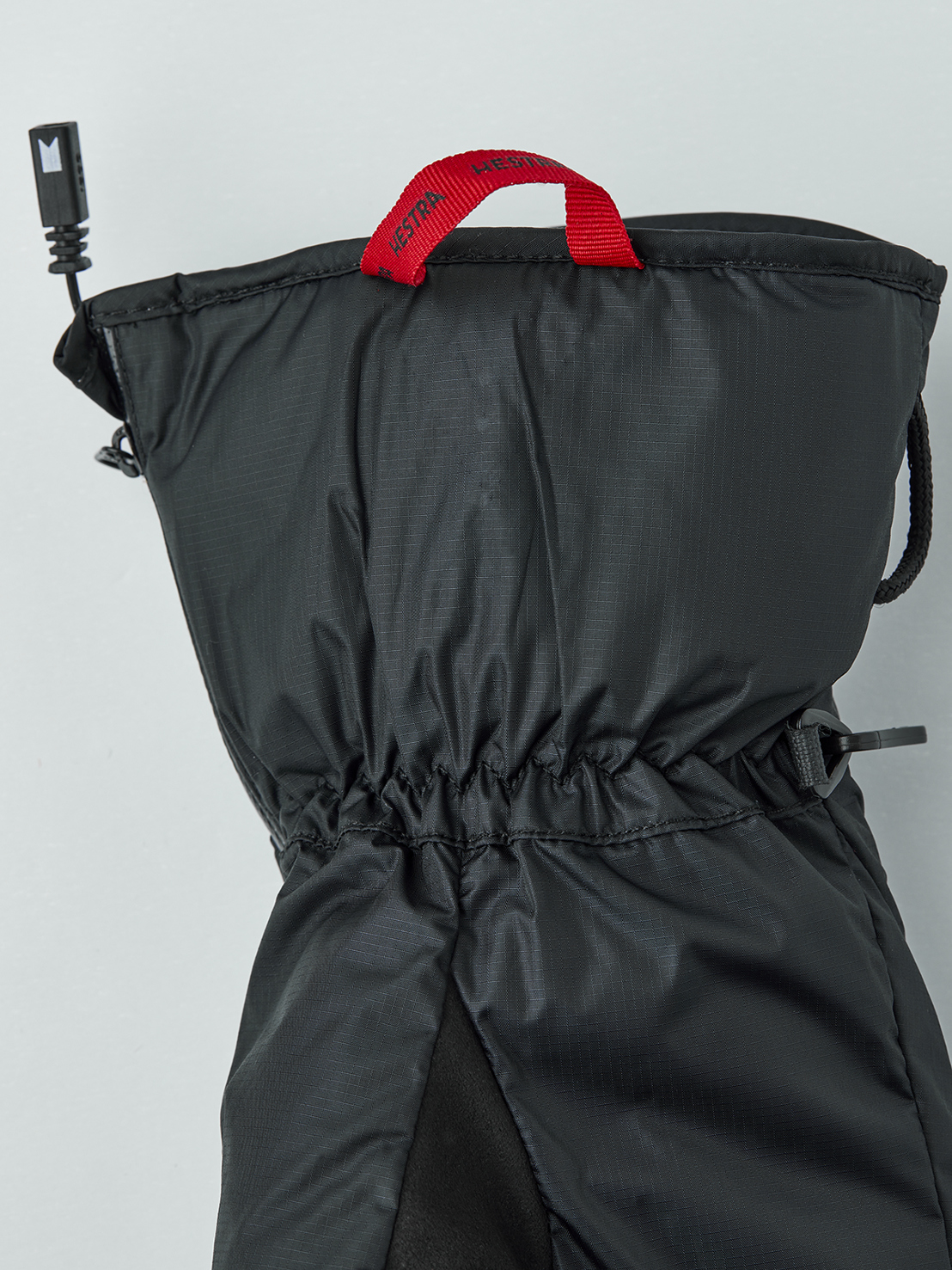 Gant intérieur chauffant noir avec dragonne rouge Hestra, cordon de serrage et câble de raccordement sur fond neutre.