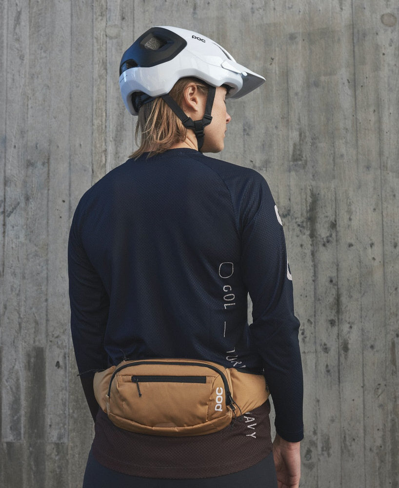 Porter un sac de hanche marron Une personne porte un sac de hanche POC Lamina brun devant un mur en béton. Elle porte également un casque et un t-shirt à manches longues.