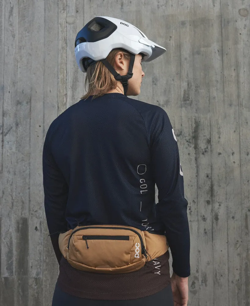 Une personne porte un sac de hanche POC Lamina brun devant un mur en béton. Elle porte également un casque et un t-shirt à manches longues.