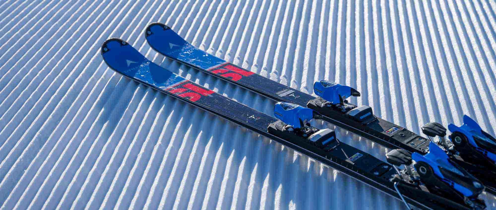 Zwei Paar Omeglass Speed Master SL Ski mit blauen Bindungen auf präparierter, geriffelter Schneeoberfläche.