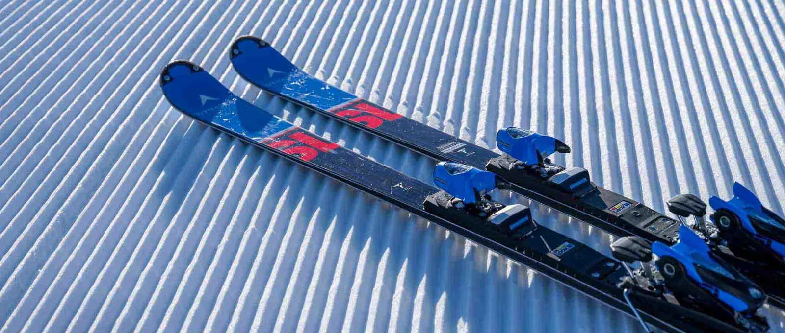 Deux paires de skis Omeglass Speed Master SL avec fixations bleues sur une surface de neige préparée et striée.