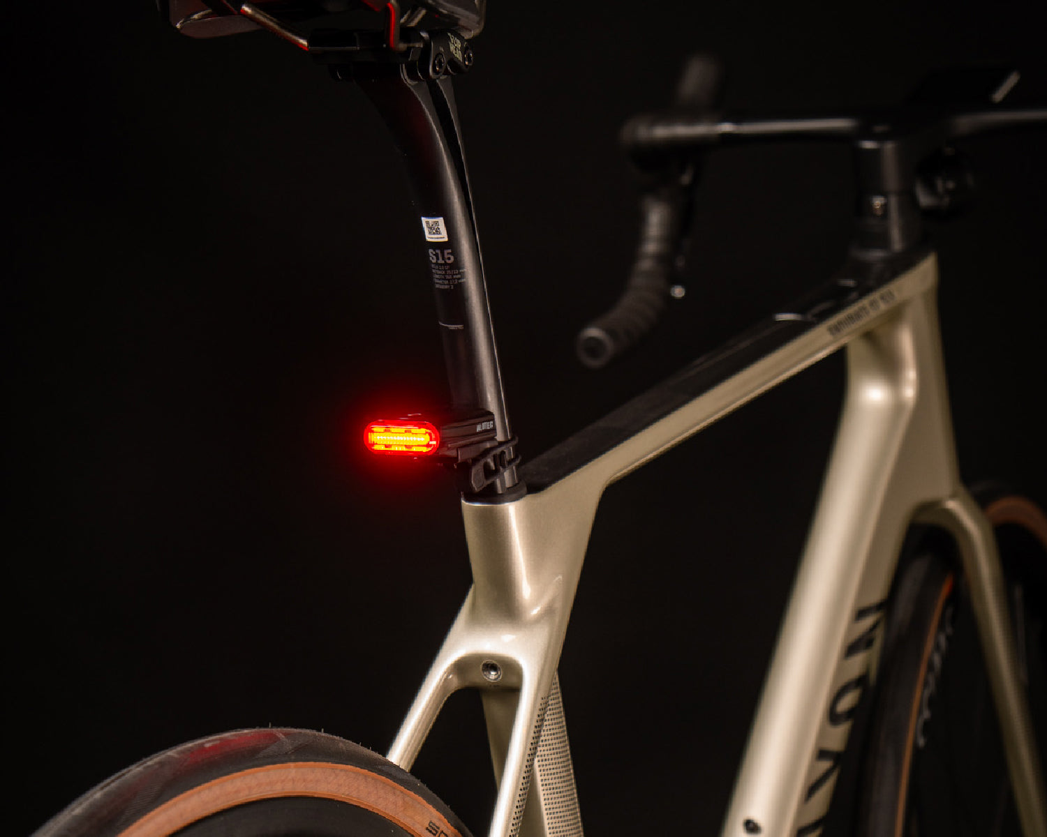 Close-up of a bicycle with red rear light switched on, seat post and parts of the frame against a black background.