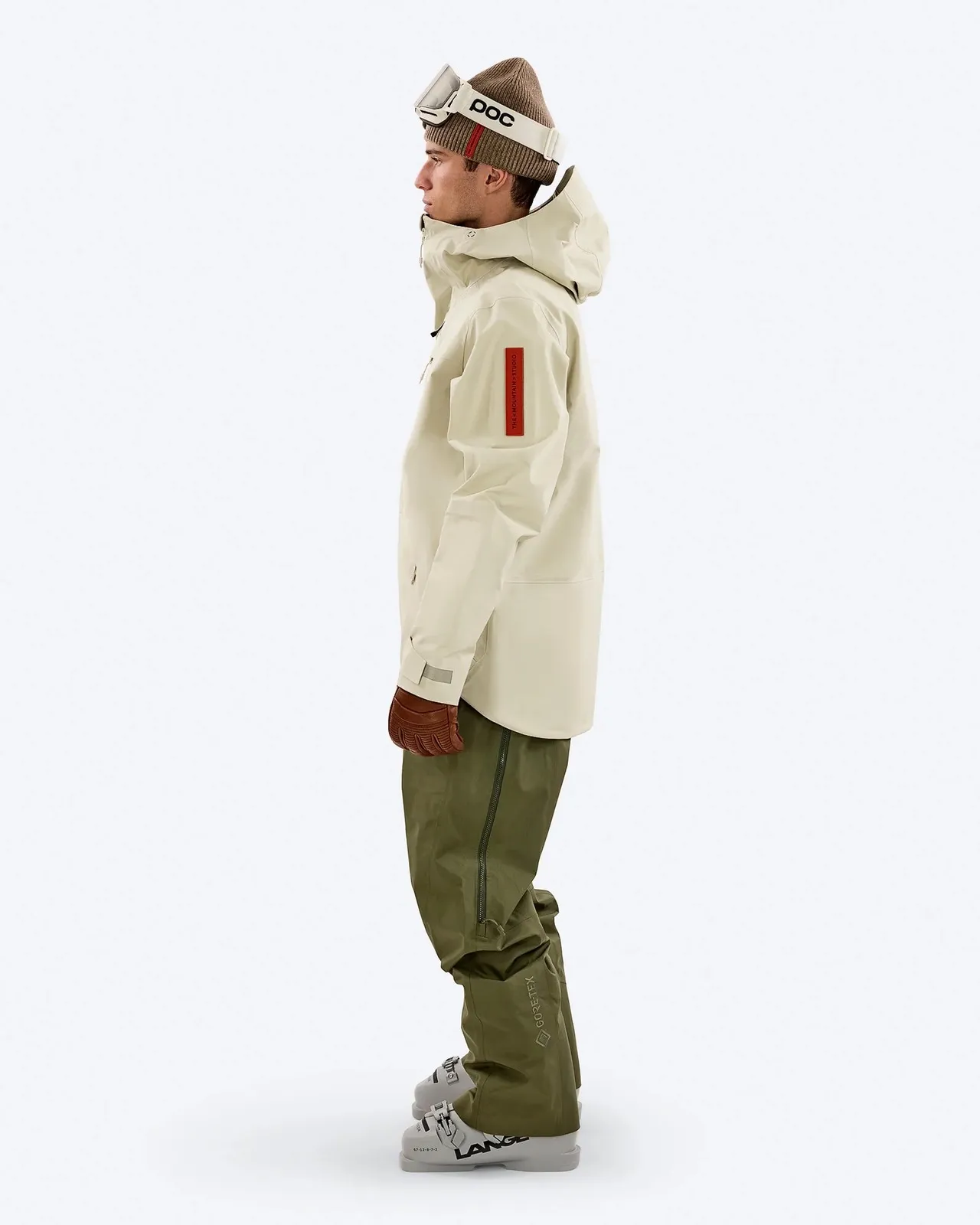 Man in ski clothing: Beige anorak with hood, green pants, ski boots and brown gloves, wearing cap and ski goggles.