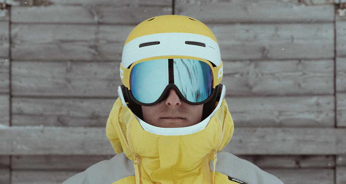 Portrait d'une personne portant un casque de ski jaune et des lunettes de ski à miroirs bleus devant un mur en bois gris. Une veste jaune est visible.