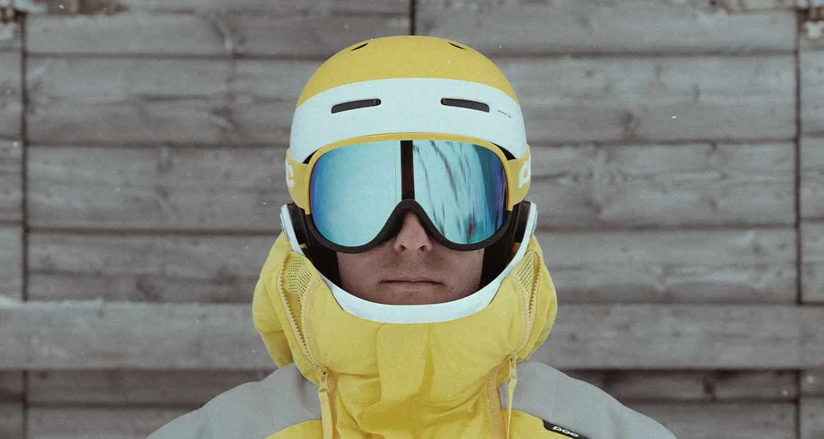Portrait of a person with a yellow ski helmet and blue mirrored ski goggles in front of a gray wooden wall. Yellow jacket is visible.