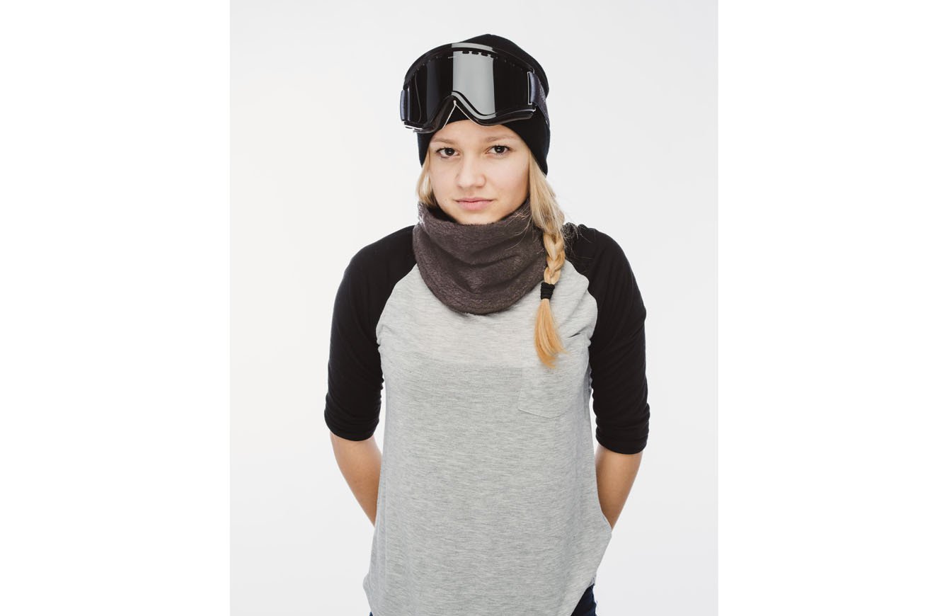 Junge Frau mit grauem Wellness Neckwarmer, Mütze und Skibrille vor weißem Hintergrund.