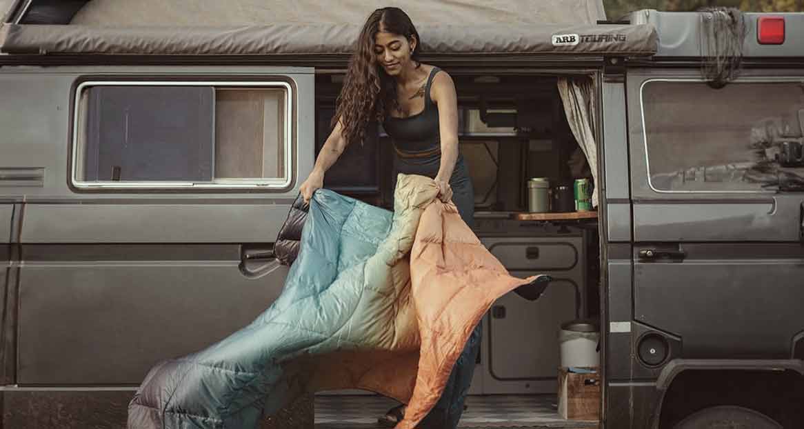 Femme avec sac de couchage devant un van. La couverture a un dégradé de couleurs allant du marron à l'orange et au bleu clair.